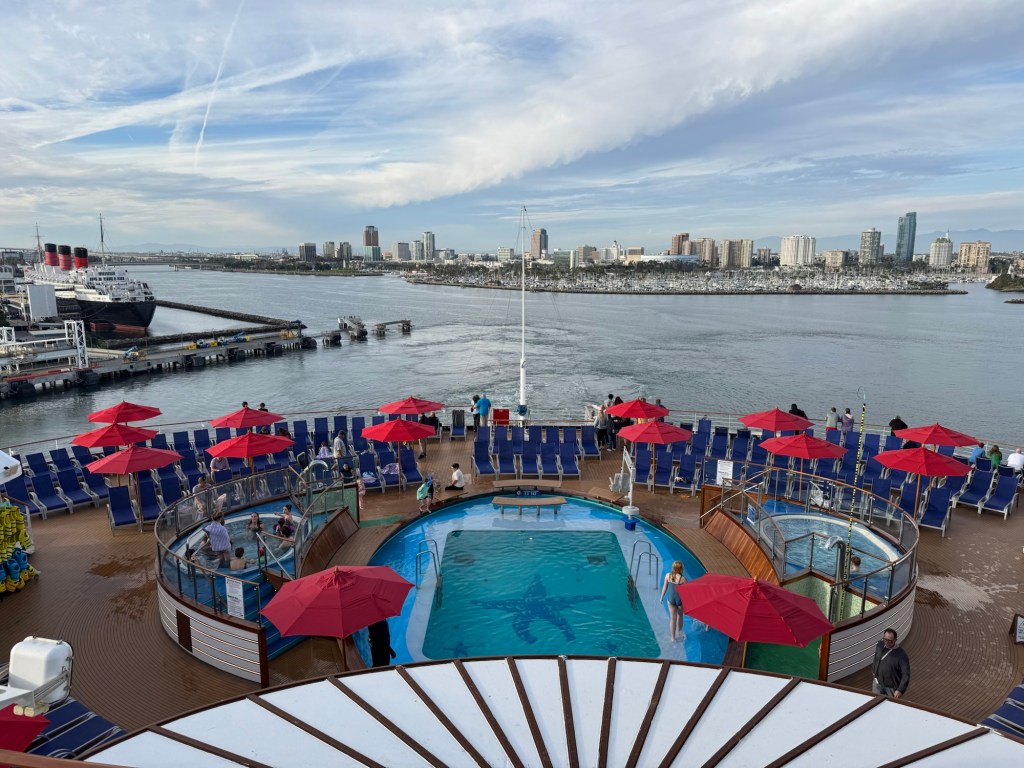 View from back of a cruise ship, with a pool and lounge chairs with a cruise port in the background